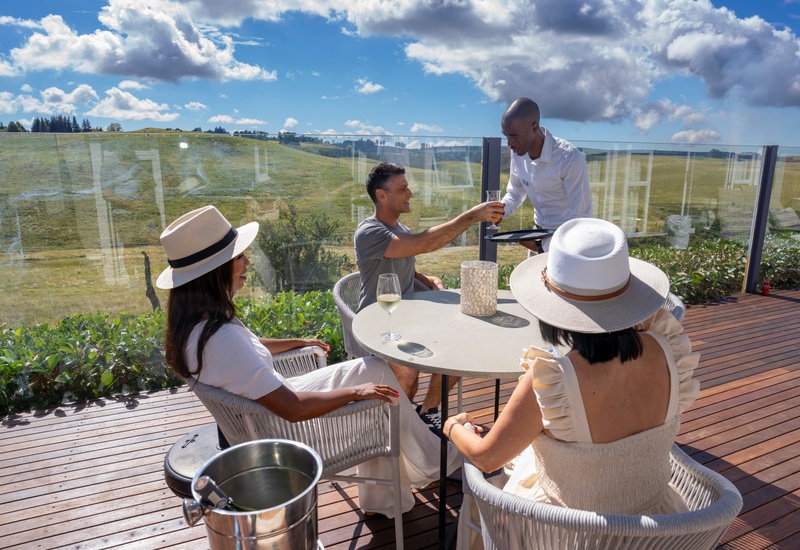 Sunny hotel terrace with glass railing, guests in sun hats enjoying wine as waiter serves drinks, rolling hills view