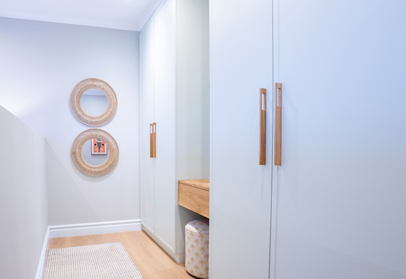 Bright hallway wardrobe with white built-in closets, wooden handles, rattan round mirrors, small wooden vanity and patterned stool.