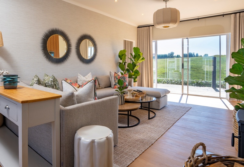Bright lounge with L-shaped sofa, coffee tables, plants and textured rug; sliding glass doors to sunlit deck and grassy view.