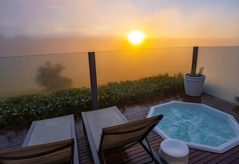 Private terrace with bubbling jacuzzi, two lounge chairs on a wet wooden deck and glass railing framing a misty sunrise.