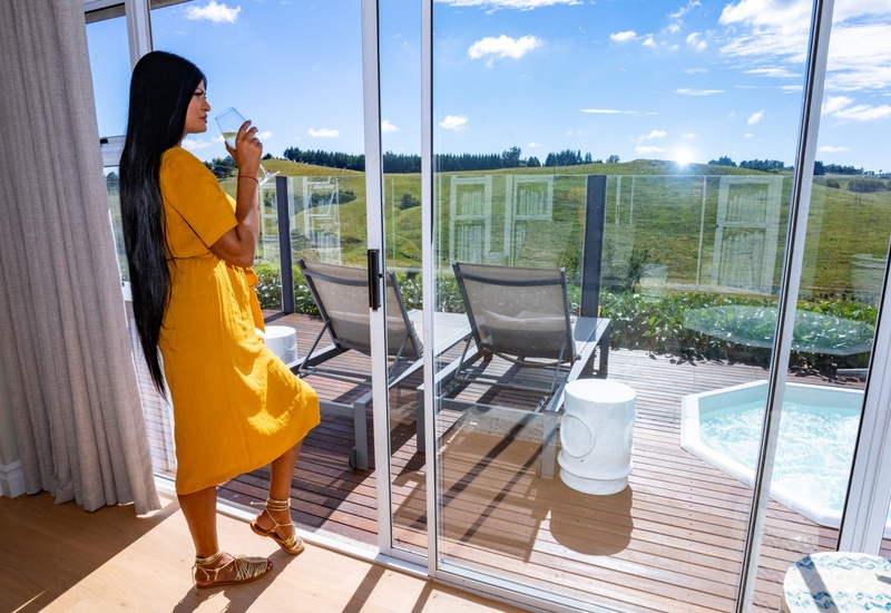 Woman in yellow dress sipping wine on a suite balcony, sunlit deck with loungers, private hot tub and rolling countryside view