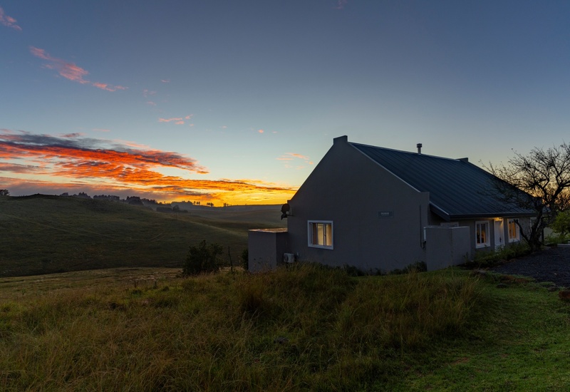 Small country cottage with metal roof and warm-lit windows beside a leafless tree, overlooking rolling grassy hills at sunrise with orange clouds.