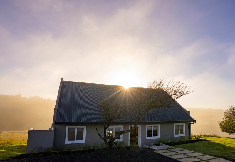 Sunrise over a country guesthouse with corrugated roof, misty rolling hills and a quiet lawn path.