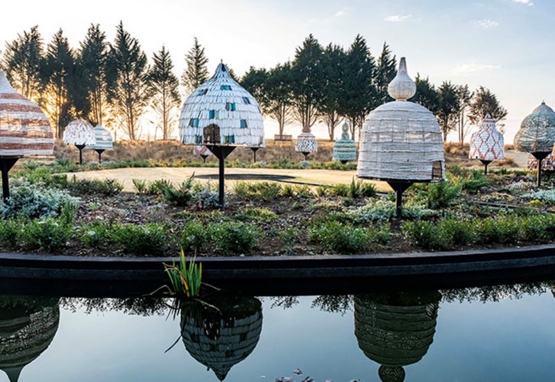 Outdoor garden of elevated beehive-style pods with colorful woven exteriors beside a reflective pond and pine trees at dawn