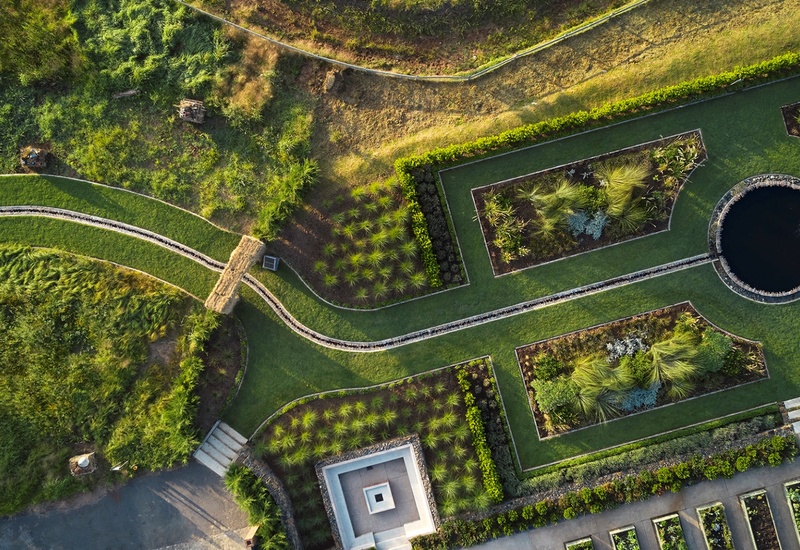 Aerial hotel gardens with geometric lawns, planted beds, winding paths and a circular reflecting pond with a small fountain.
