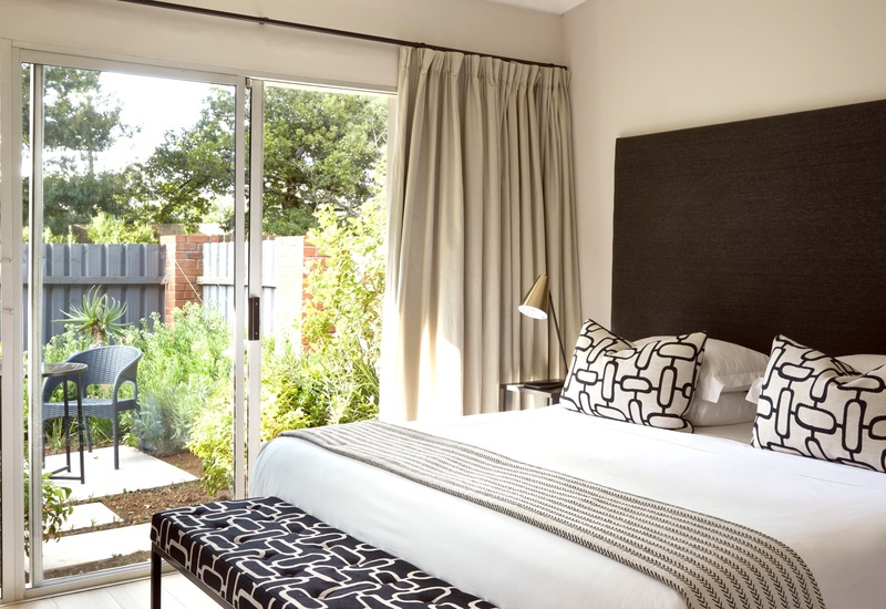 Bright king bedroom with large sliding glass door opening to private garden patio, patterned cushions, bench and bedside lamp.