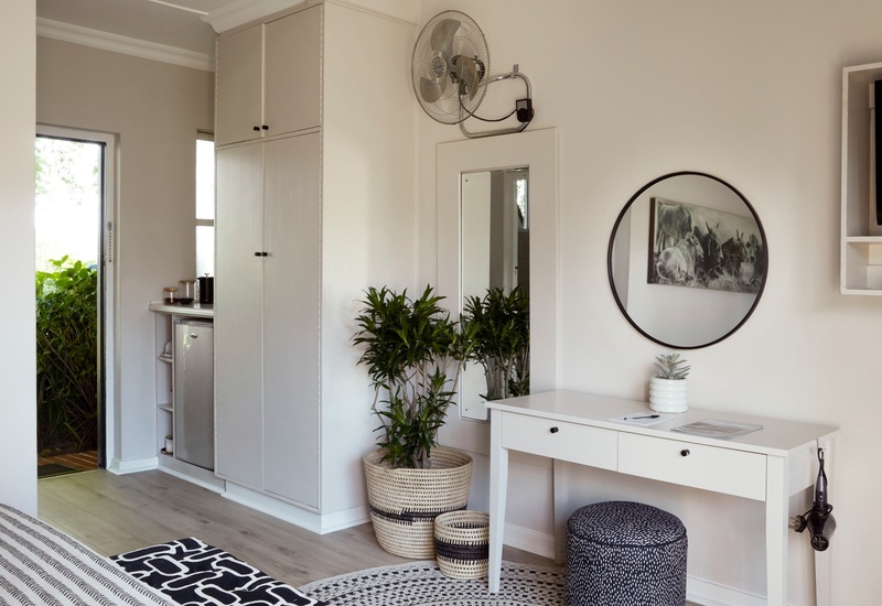 Bright airy guest room with white furnishings, built-in wardrobe and mini-fridge, desk with round mirror, woven plant baskets and garden doorway view