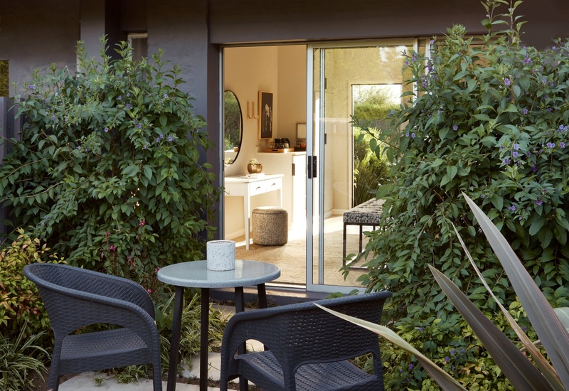 Private patio with two wicker chairs and round table, sliding glass door opens to a bright room with vanity and pouf, surrounded by greenery.