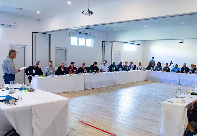 Bright modern meeting room with U-shaped white-clothed tables, overhead projector, speaker presenting to seated guests with water jugs.