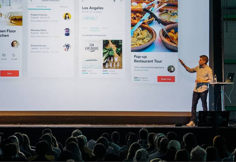 Speaker on stage beside giant projected booking screens showing loft, Los Angeles experiences and pop-up restaurant tour.