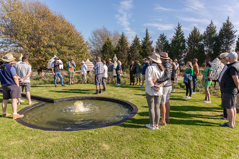 Guests on a guided garden tour around a circular fountain on a manicured lawn, surrounded by sculptural garden huts and mature trees.