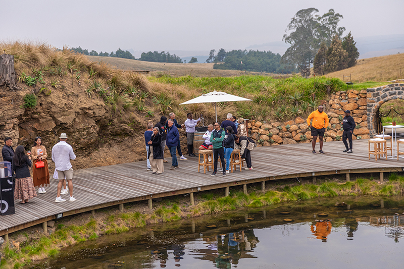 Countryside waterfront terrace with wooden deck, guests mingling under umbrella, stone arch, rolling hills views.