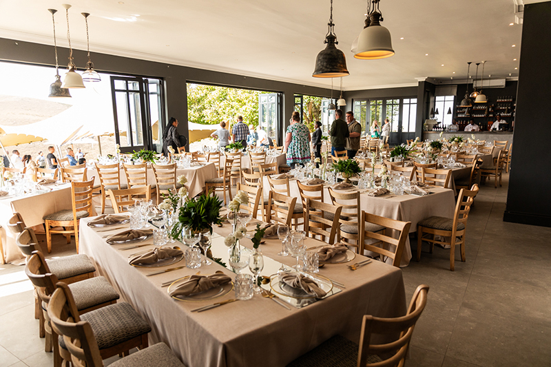 Elegant dining room set for reception: long tables with beige linens, glassware and flowers; guests mingle by open terrace.