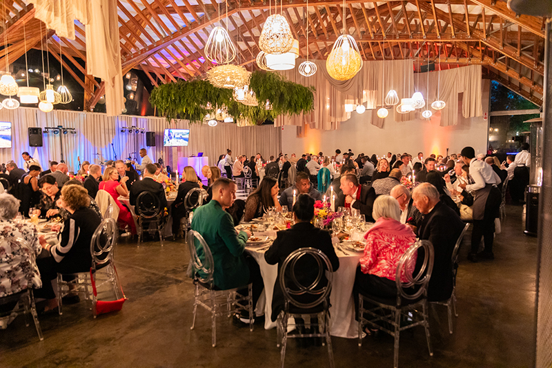 Candlelit banquet in a rustic hall with vaulted wooden ceiling, pendant lights, hanging greenery and guests dining.
