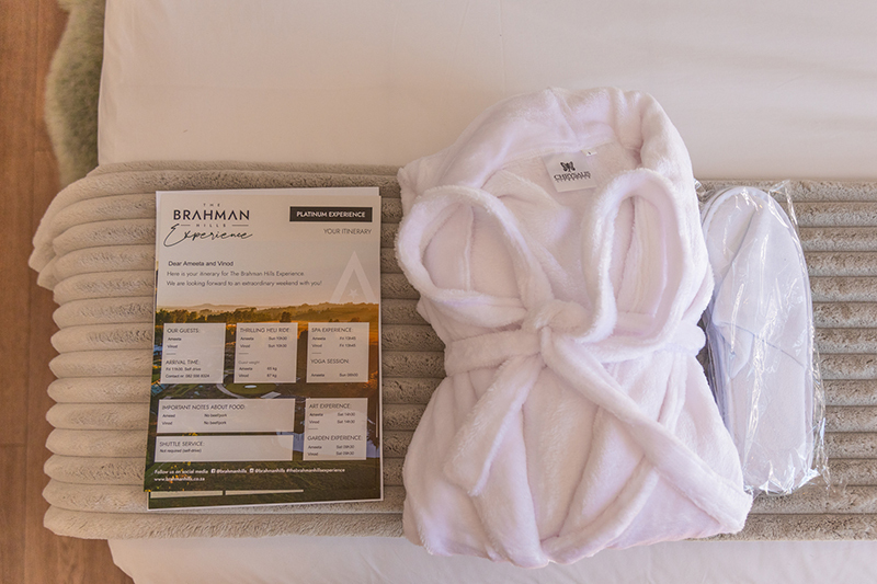 Plush white bathrobe tied on bed with packaged slippers and a welcome itinerary card on a textured folded throw.