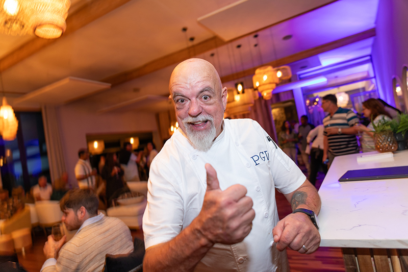 Smiling chef in white jacket giving thumbs-up in lively hotel lounge, warm chandeliers, purple uplighting and mingling crowd