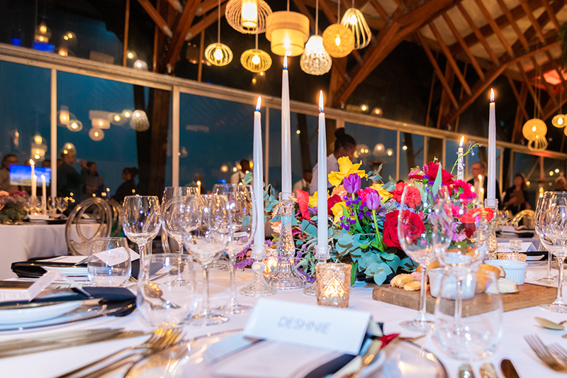 Candlelit banquet table with colorful floral centerpiece, glassware and candles under warm pendant lights in beamed hall