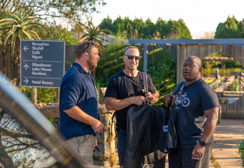 Three male guests chatting near hotel directional sign in sunlit gardens, carrying luggage and garment bags at arrival.