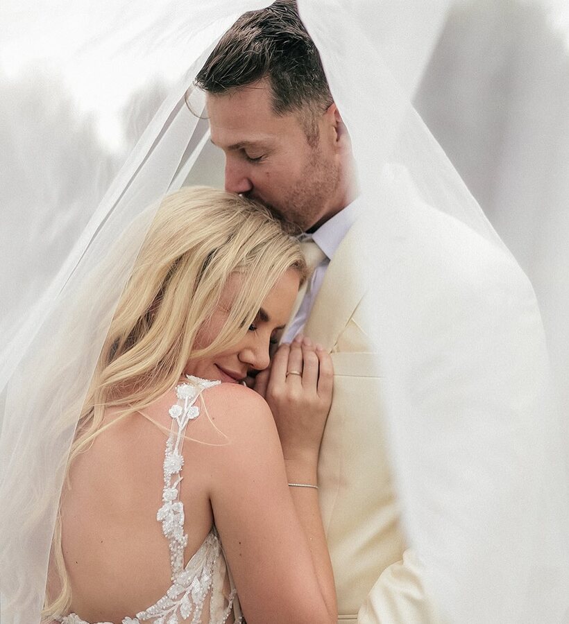 Bride in beaded backless gown and groom in cream suit embrace under veil as he kisses her forehead — wedding portrait.