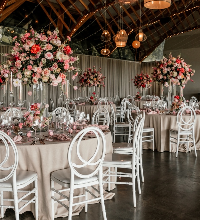 Banquet hall set for wedding with round tables, blush linens, tall pink floral centerpieces, white chairs and warm hanging lights.