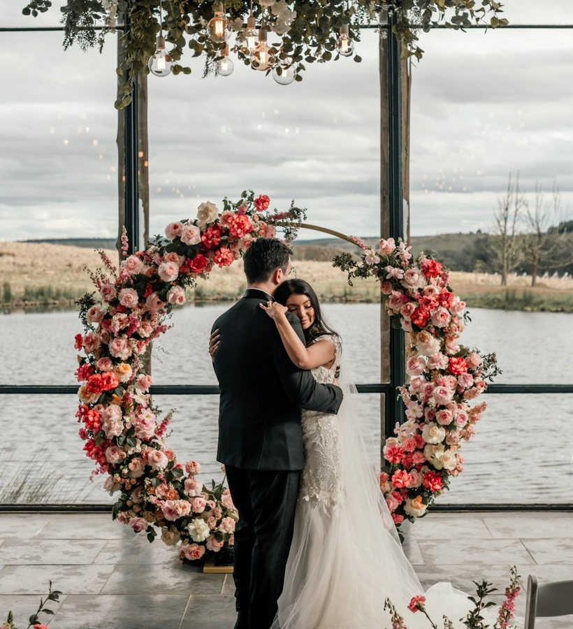 Bride and groom embrace beneath a lush floral arch and hanging bulbs in a lakeside wedding venue with floor-to-ceiling windows.