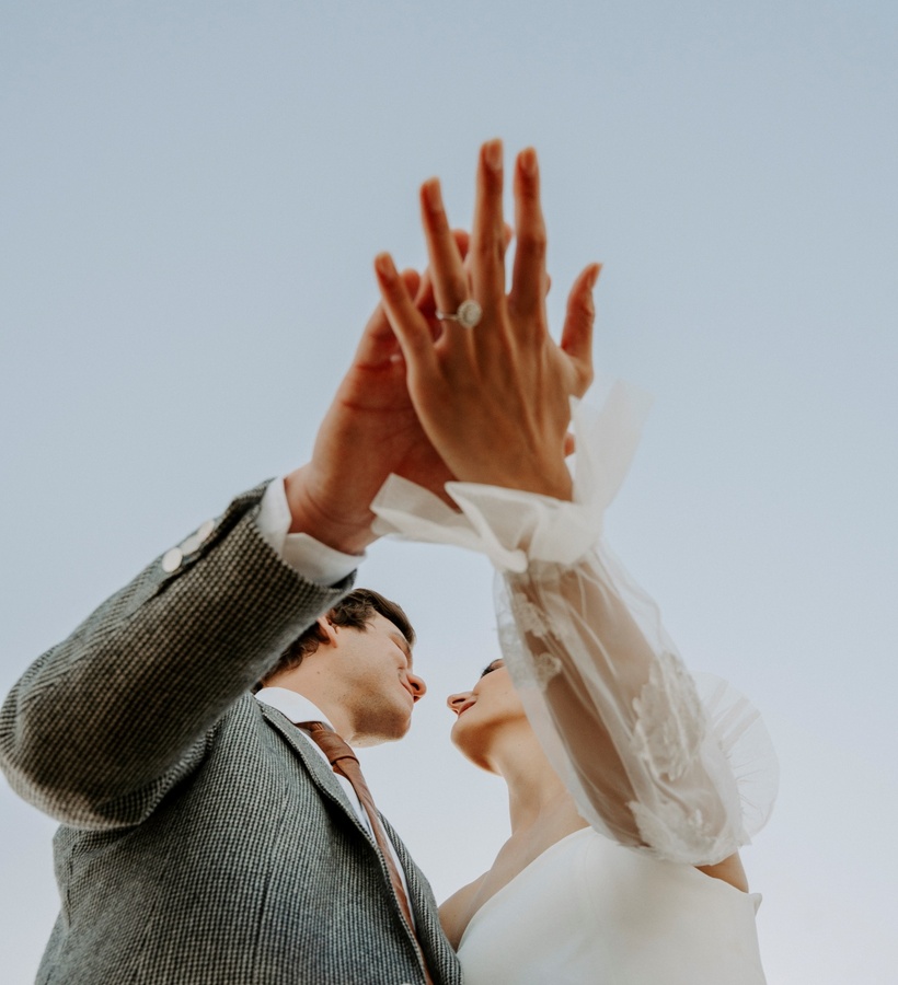 Newlyweds in elegant wedding attire raise clasped hands to show a ring against a pale sky, a romantic hotel wedding moment