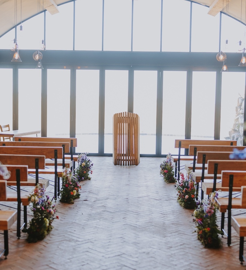 Stone-walled chapel with arched glass wall, wooden benches, floral-lined aisle, central wooden lectern and warm hanging pendant lights
