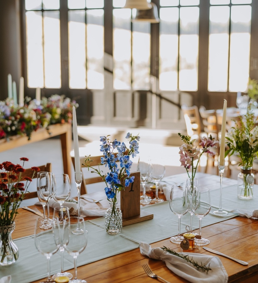 Sunlit dining room with wooden banquet tables set for celebration, glassware, taper candles and fresh floral centerpieces.