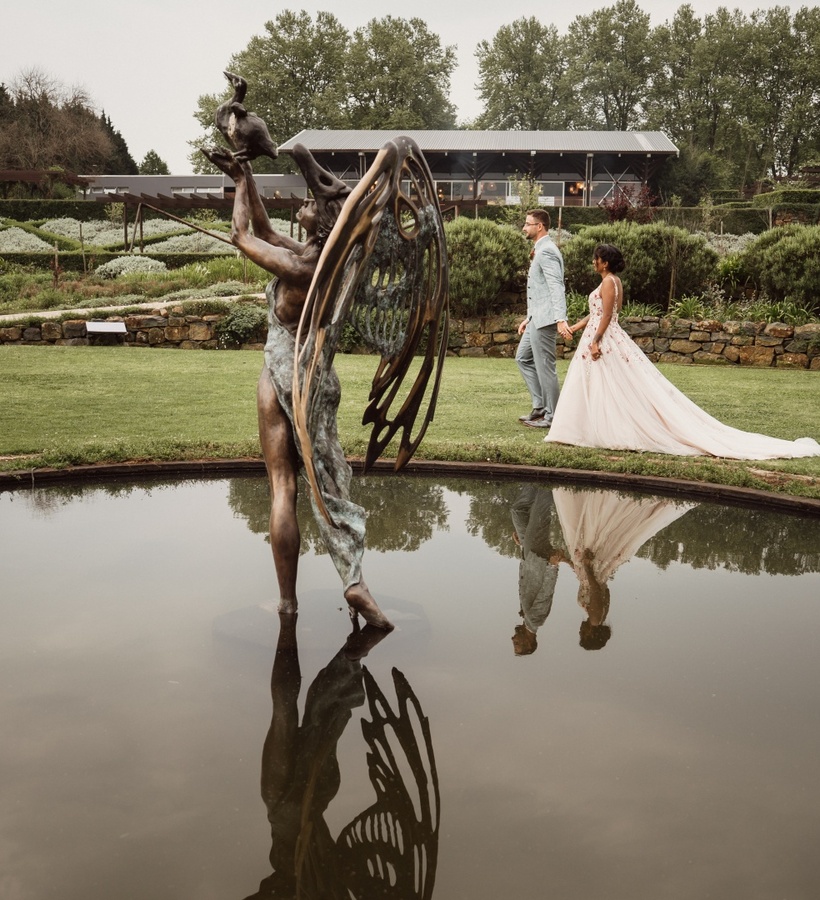 Bronze winged statue reflected in a pond, bride in flowing gown and groom walking hand-in-hand across manicured garden wedding venue.