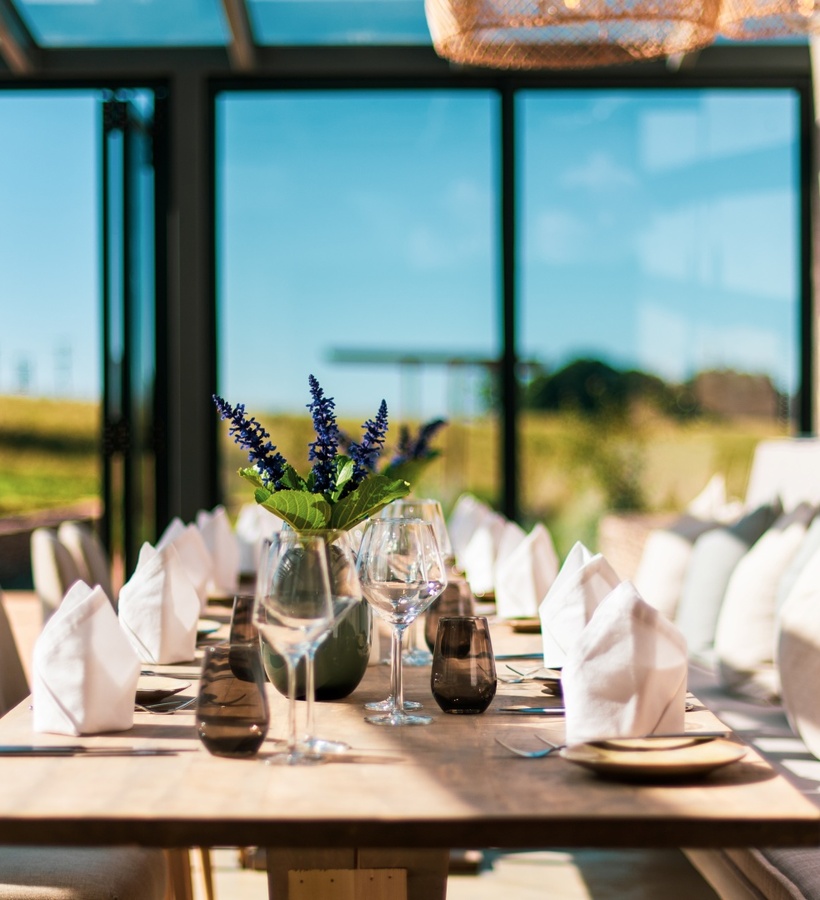 Sunlit dining table with folded napkins, wine glasses and lavender centerpiece by floor-to-ceiling windows with countryside view.