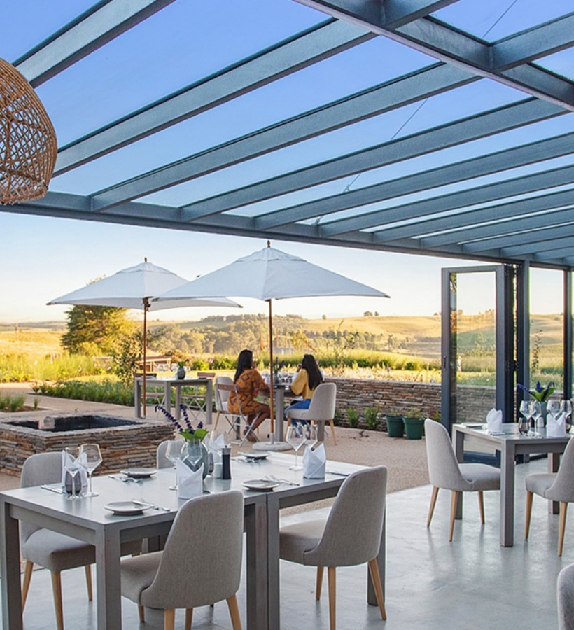 Open-air hotel restaurant with glass pergola, woven pendant lights, neatly set tables and umbrellas overlooking rolling countryside at sunset.