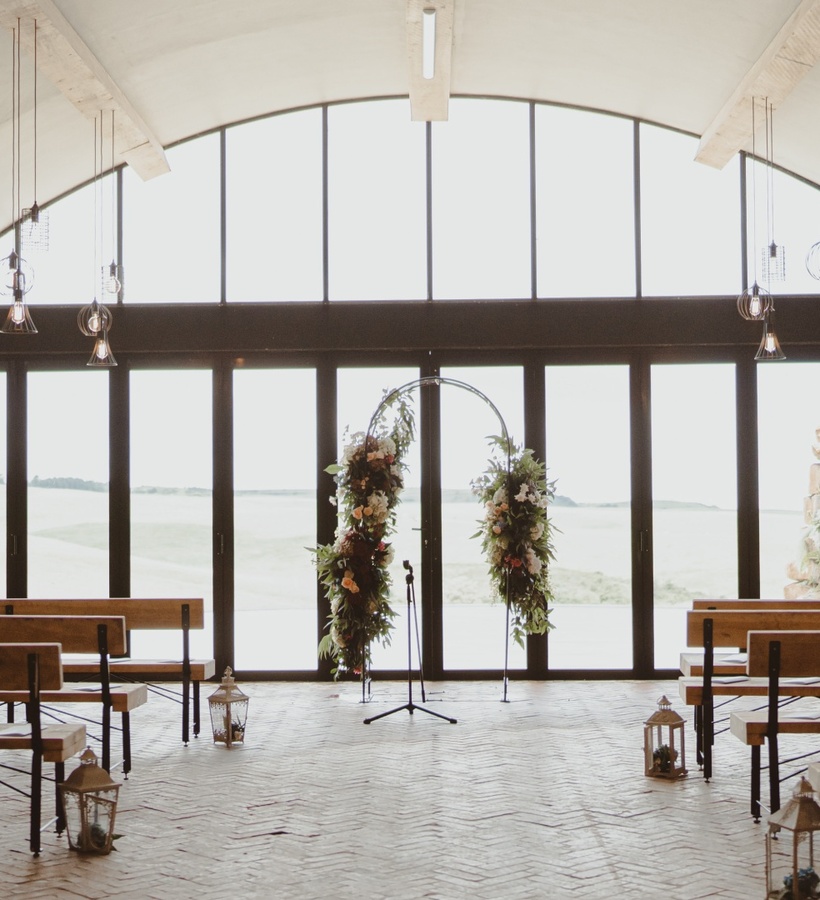 Stone-walled ceremony room with wooden benches, hanging pendant lights, floral arch and arched windows overlooking fields