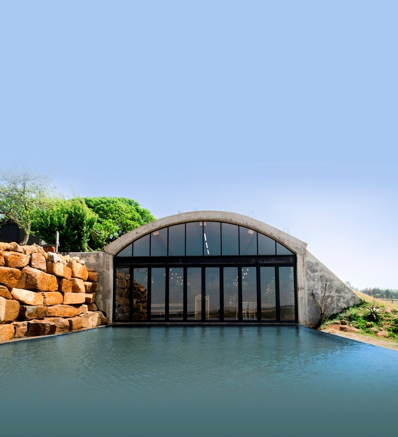 Arched concrete building with floor-to-ceiling glass doors opening onto a reflecting pool, stacked stone wall and green landscaping.