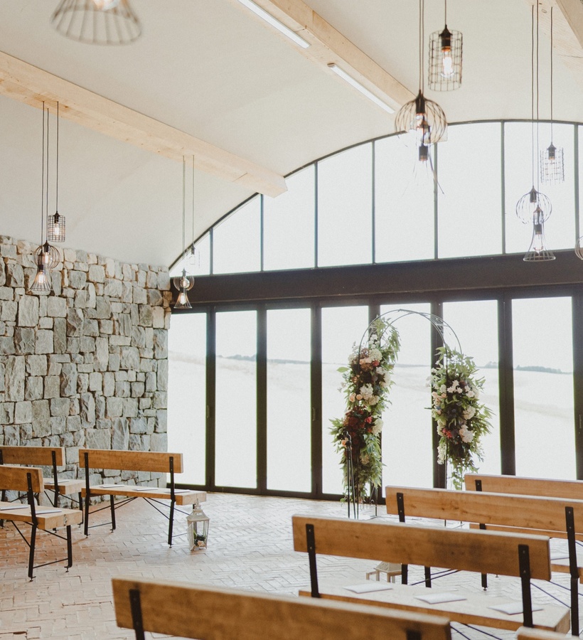 Bright ceremony hall with arched tall windows, stone wall, pendant lights, wooden benches and a floral arch.