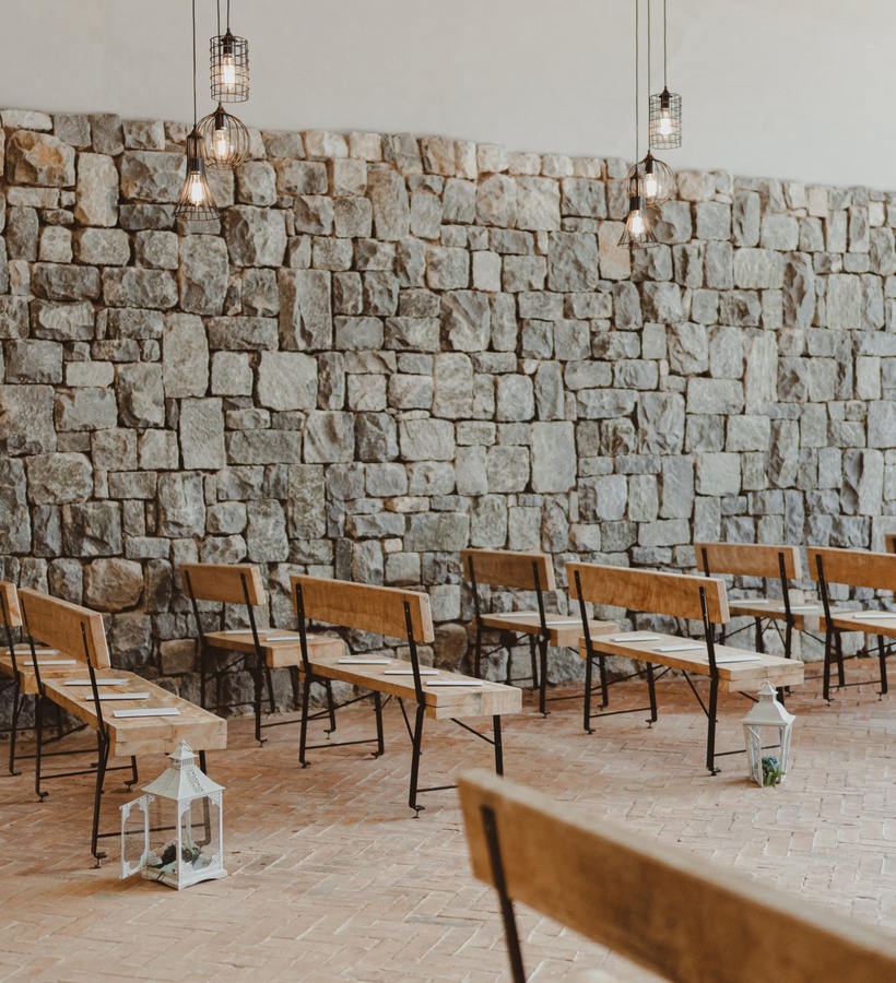 Rustic event space with stone wall, rows of wooden benches, hanging cage pendant lights and white lanterns lining the aisle