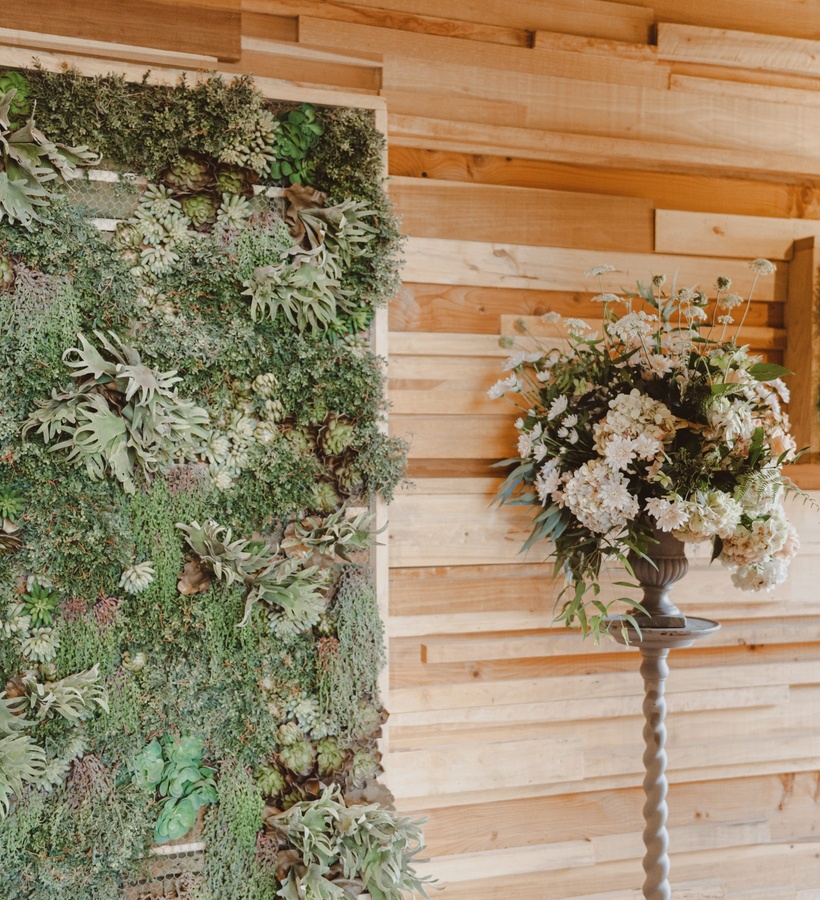 Vertical succulent green wall beside a wooden-paneled backdrop and urn of soft white flowers on a tall pedestal