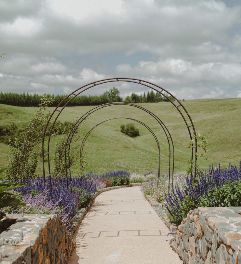 Garden walkway framed by metal arched trellises, stone walls and beds of purple flowers with rolling green hills.
