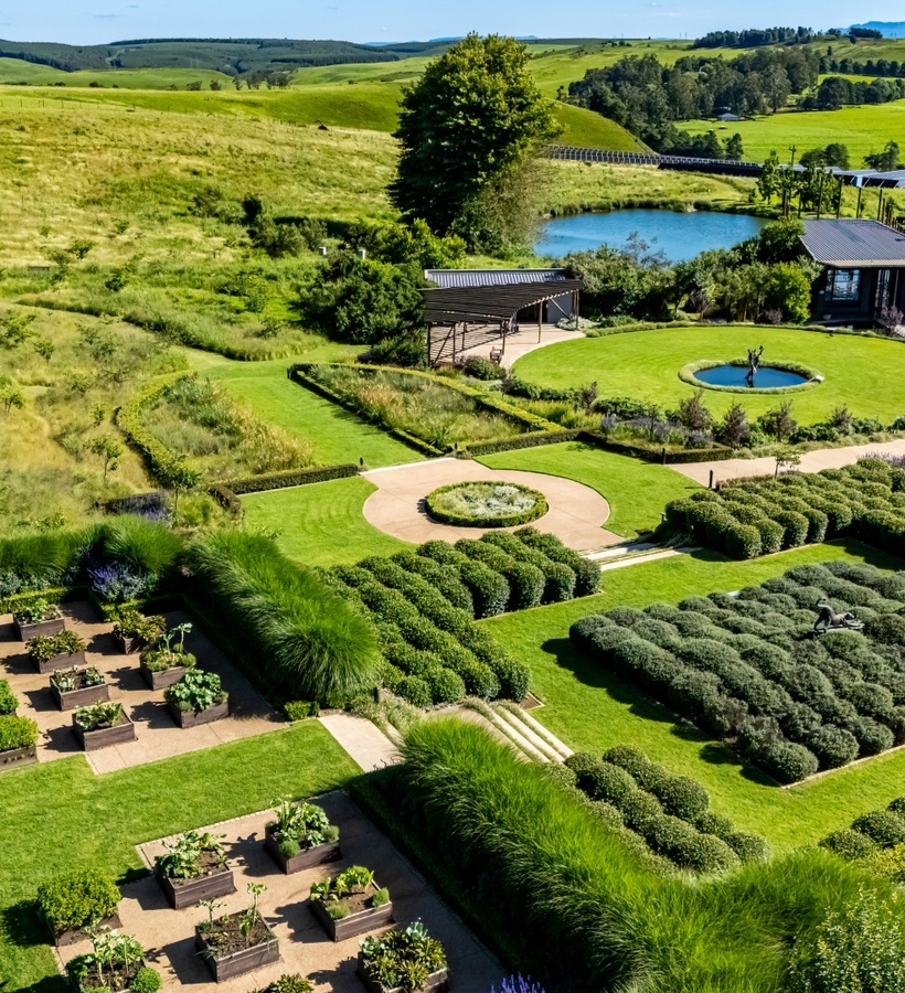 Manicured hotel gardens with raised kitchen beds, geometric hedges and a circular lawn fountain beside a pavilion and pond.