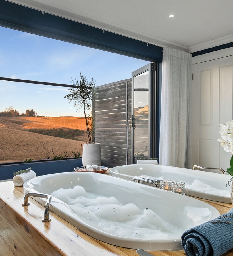 Dual soaking tubs with bubbles on a wooden platform, open terrace to golden hillside sunset, orchid and rolled towels.