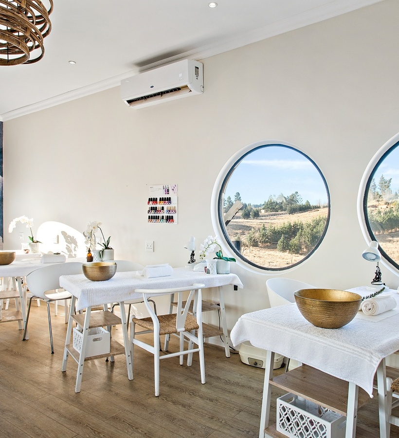 Bright spa manicure room with white treatment tables, gold bowls, orchids, rattan pendant lights and round windows framing rolling countryside views.