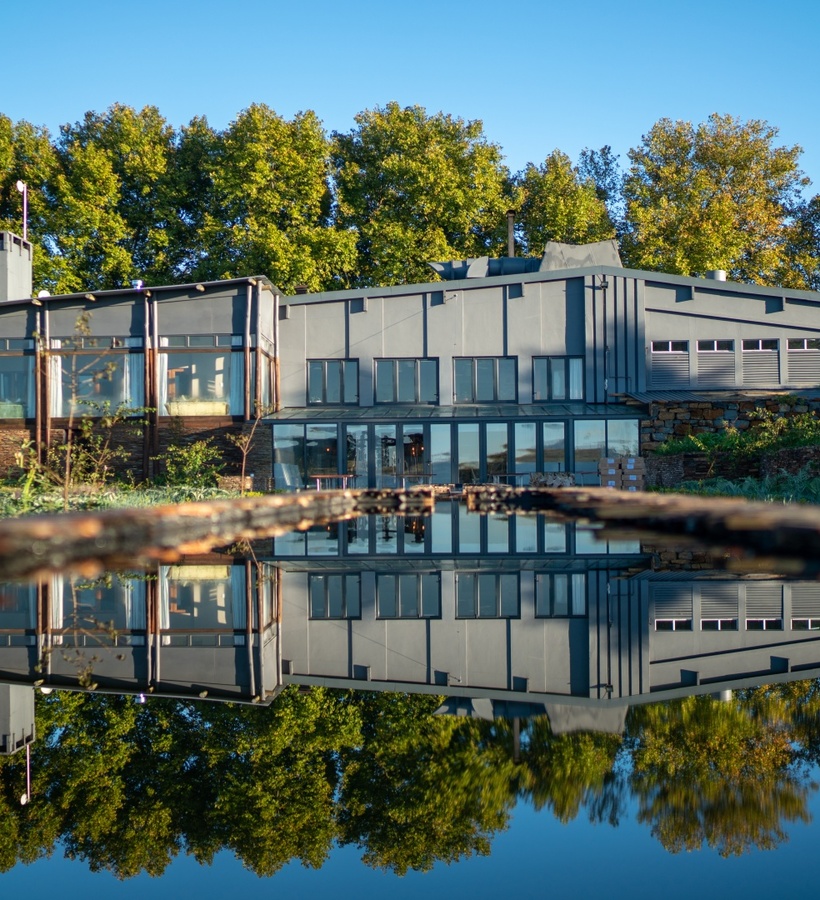 Modern lakeside lodge with floor-to-ceiling windows, wooden decks and stone terraces reflected in a calm pool, framed by tall green trees.