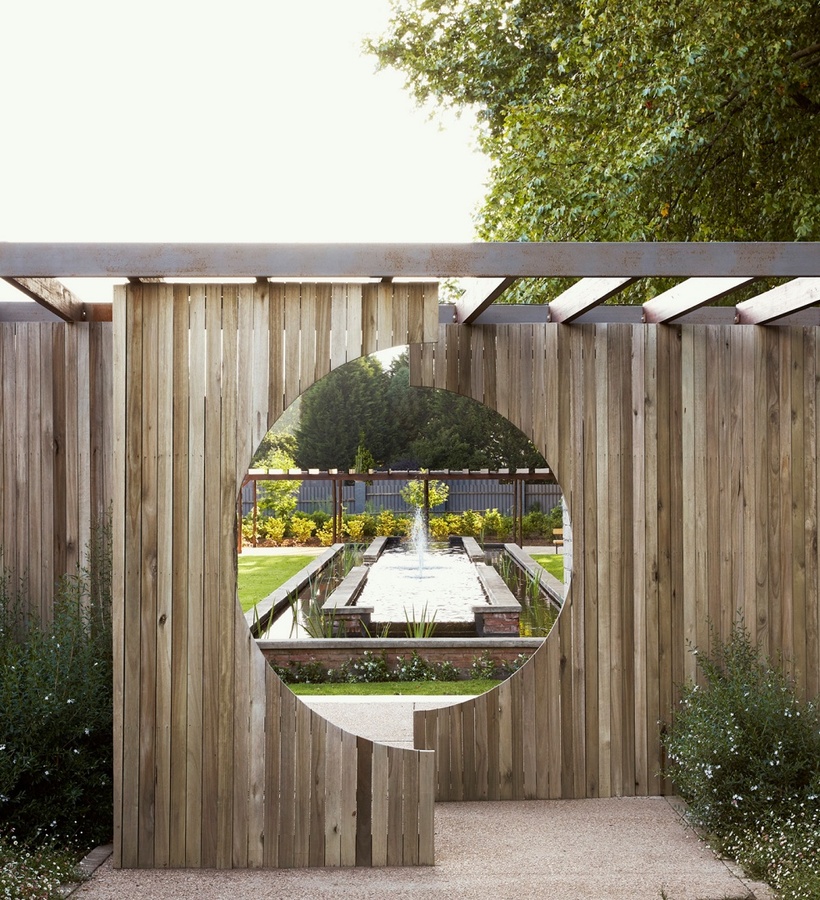 Wooden pergola and privacy screen with circular cutout framing a long reflecting pool and fountain amid manicured gardens.