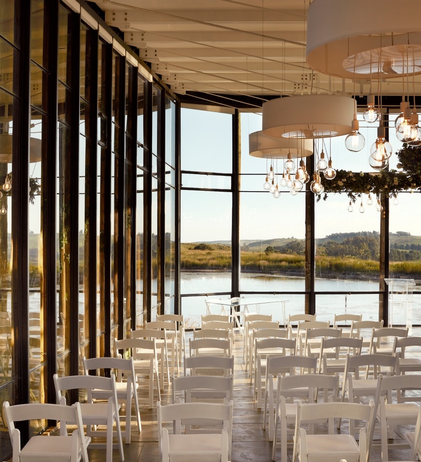 Glass-walled lakeside event space set for a ceremony with white folding chairs, hanging bulbs and floral wreaths.