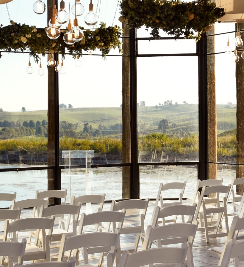 Glass-walled ceremony space with white folding chairs, hanging bulb lights, greenery and serene meadow and pond views.