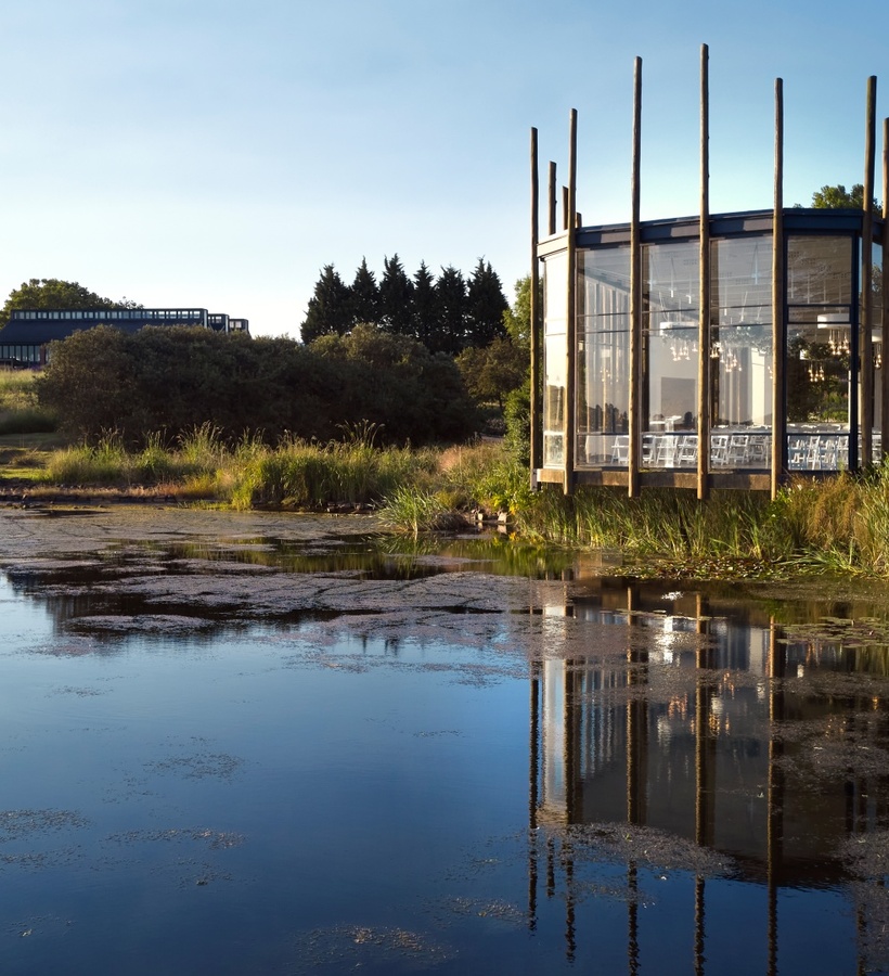 Glass lakeside pavilion on wooden stilts with chandeliers and dining chairs, reflecting on a calm pond by grassy grounds.