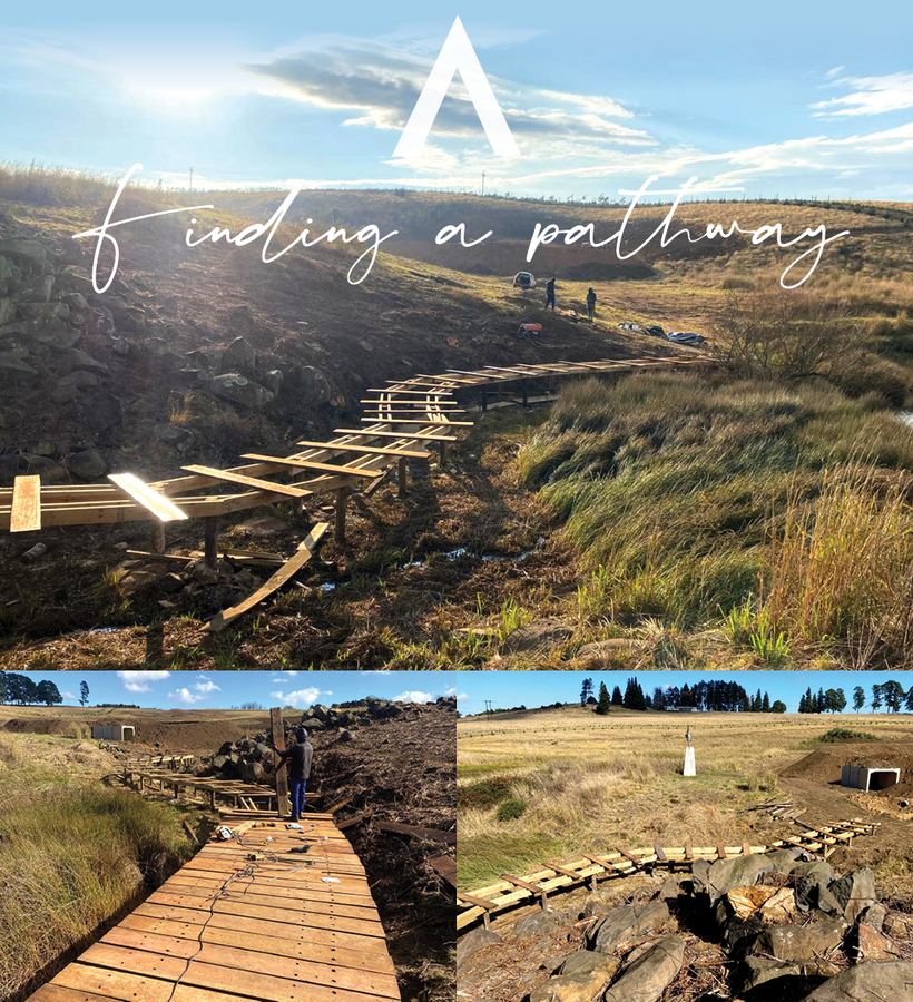 Sunlit wooden boardwalk trail under construction winding through grassy wetlands to scenic rolling hills; workers and tools visible.