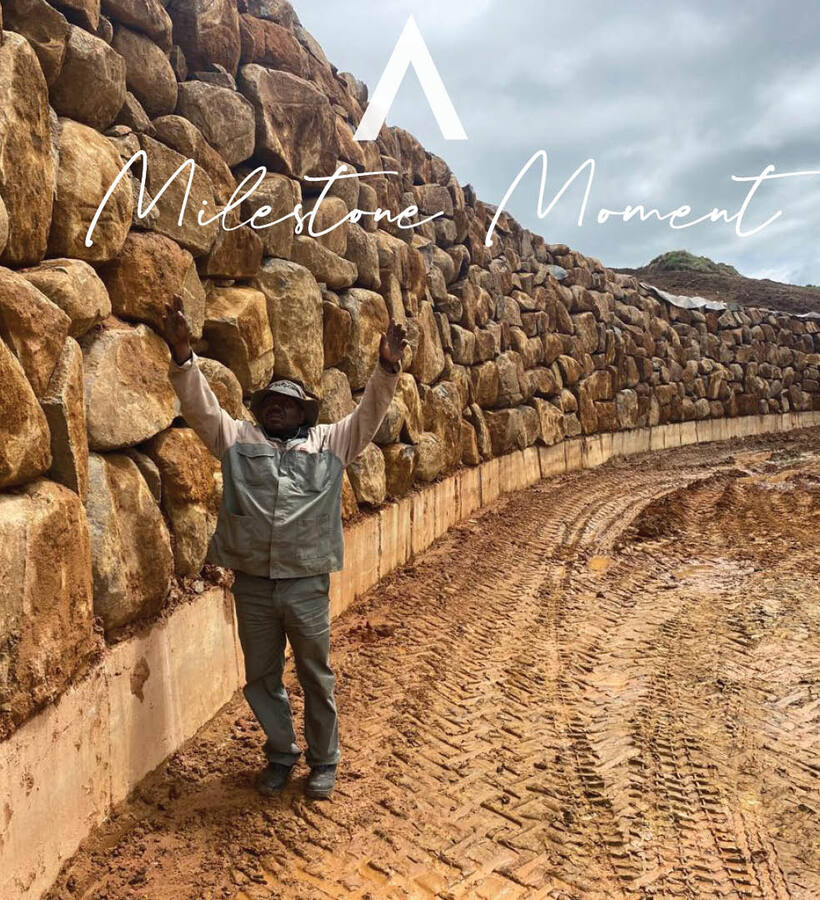 Site worker raising arms beside massive curved stone retaining wall and muddy vehicle tracks beneath cloudy sky