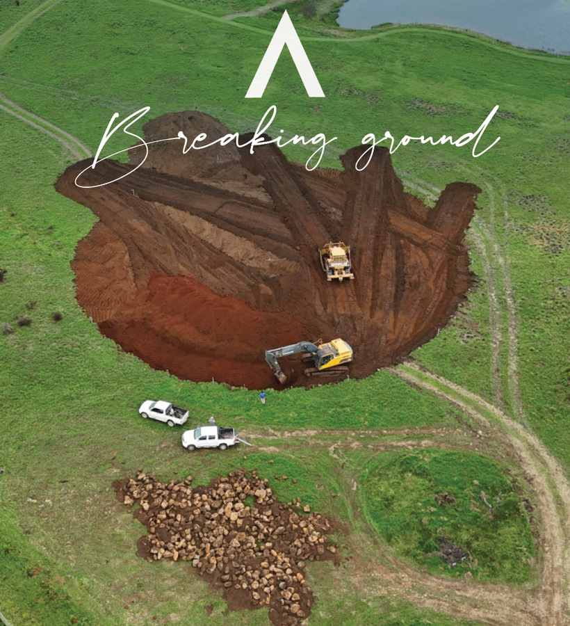 Aerial view of circular construction pit in green pasture with excavator, bulldozer, two white trucks, rock pile and 'Breaking ground' overlay