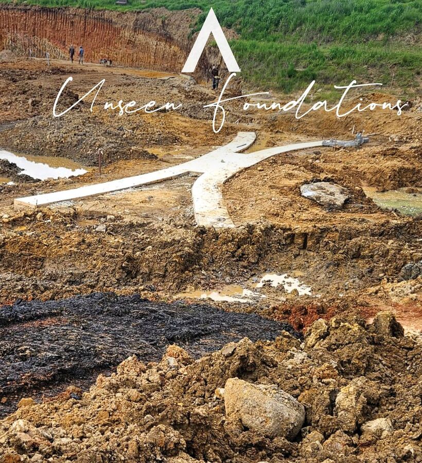 Concrete foundation cross and pathways in a muddy construction pit with two workers and grassy slope