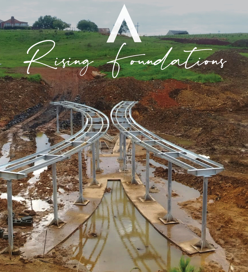 Steel elevated track foundations over a muddy construction pit with pooled water, green hillside and distant houses; 'Rising foundations' overlay.